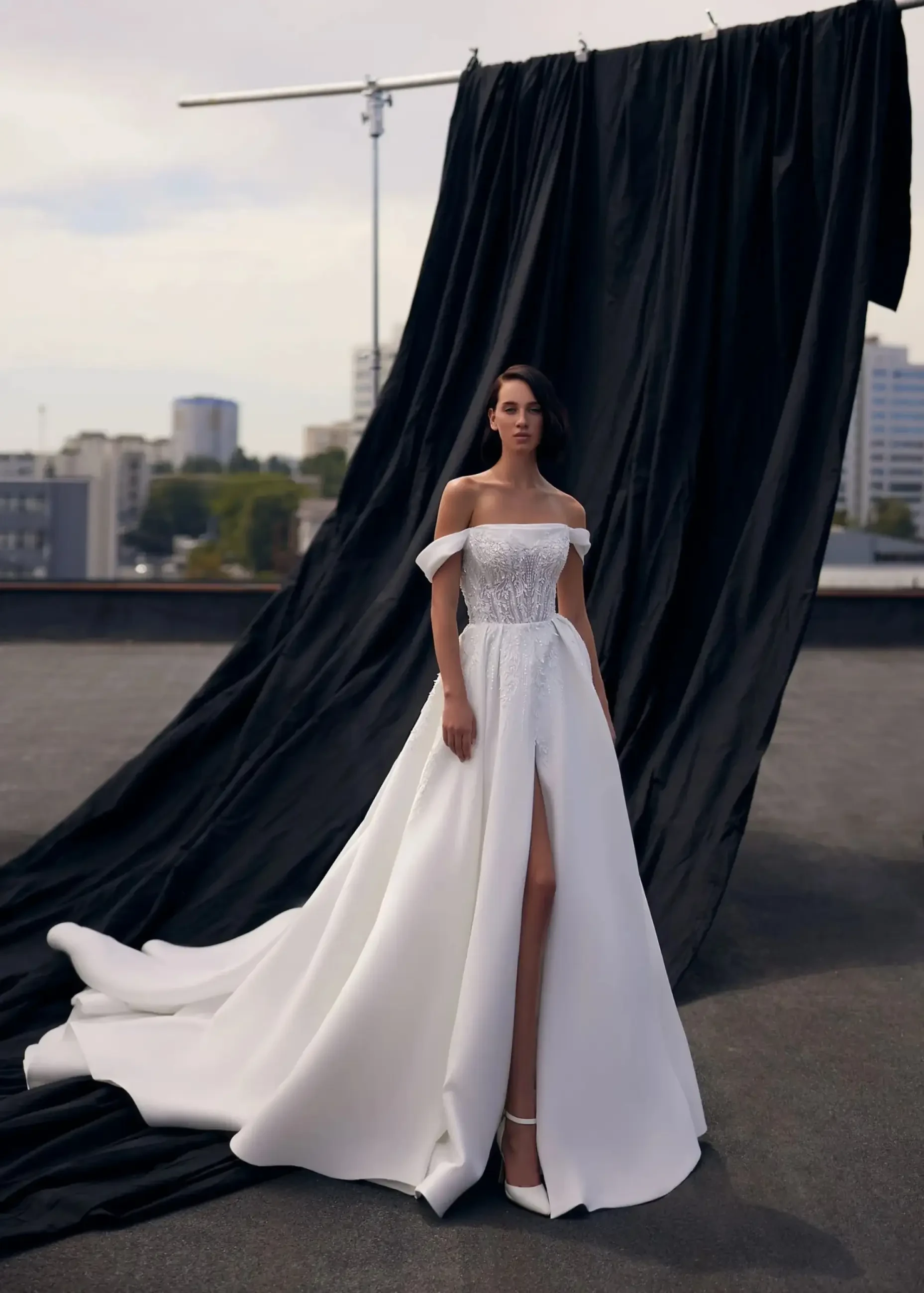 A woman in an elegant off-shoulder white gown stands confidently on a rooftop. A long black fabric drapes behind her. The scene is urban and serene.
