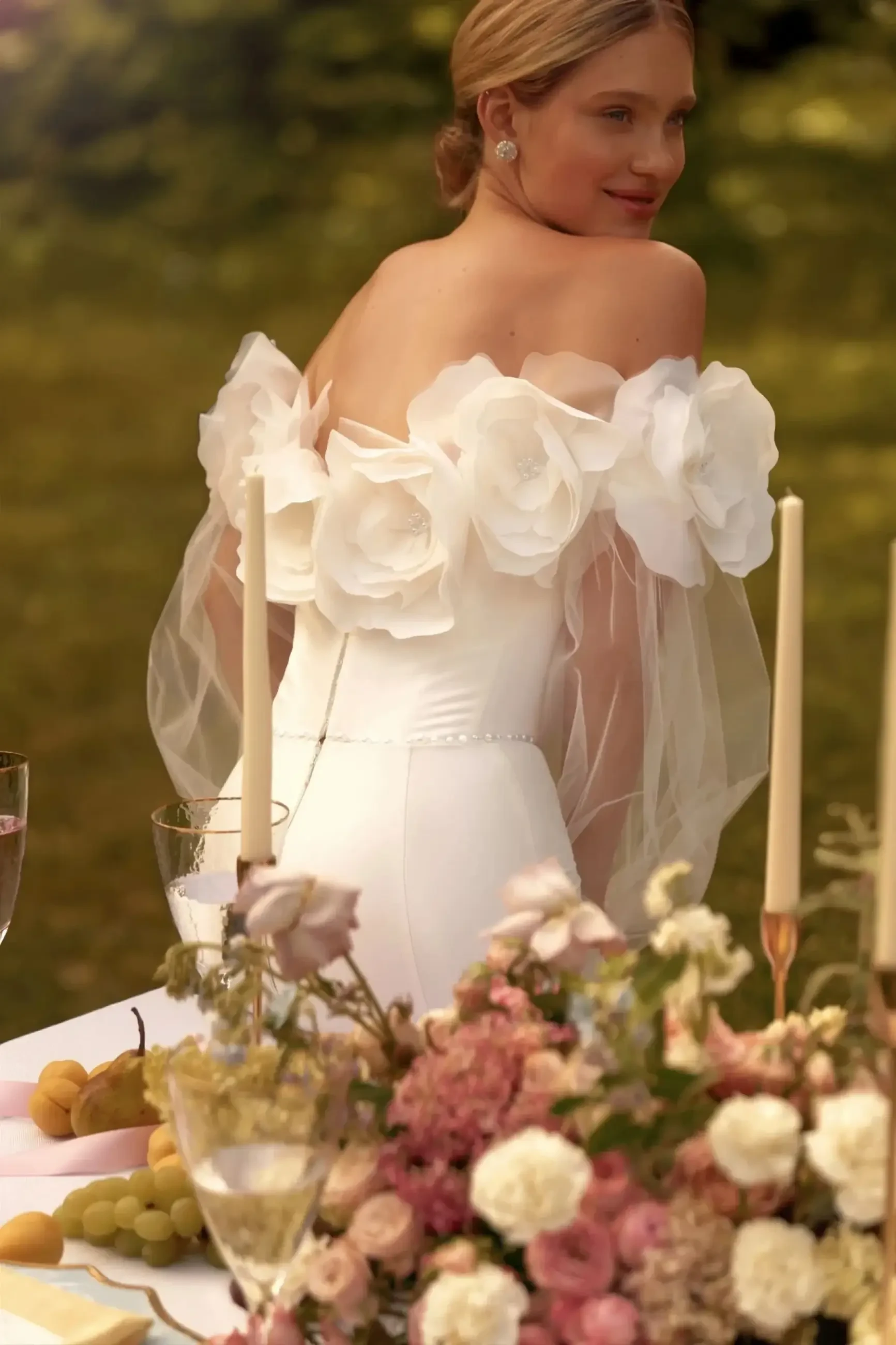Model is wearing a sculpted mermaid wedding dress with 3D floral appliqués, a curly neckline, tulle puff sleeves, and a front slit.