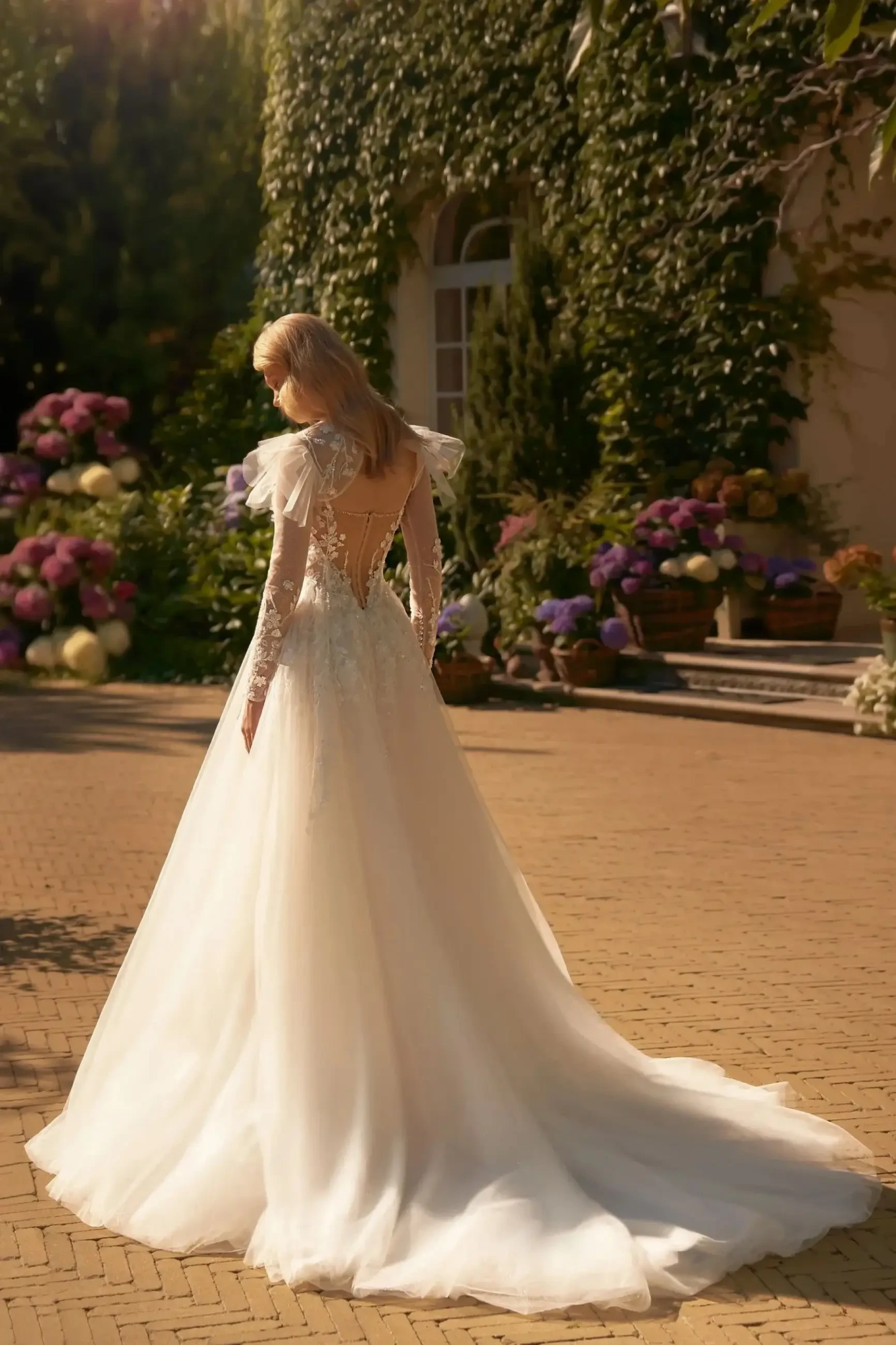 Model is wearing a soft A-line wedding gown with beaded straps, a pleated corset bodice, floral appliqués, and a layered glitter tulle skirt with a removable bolero.
