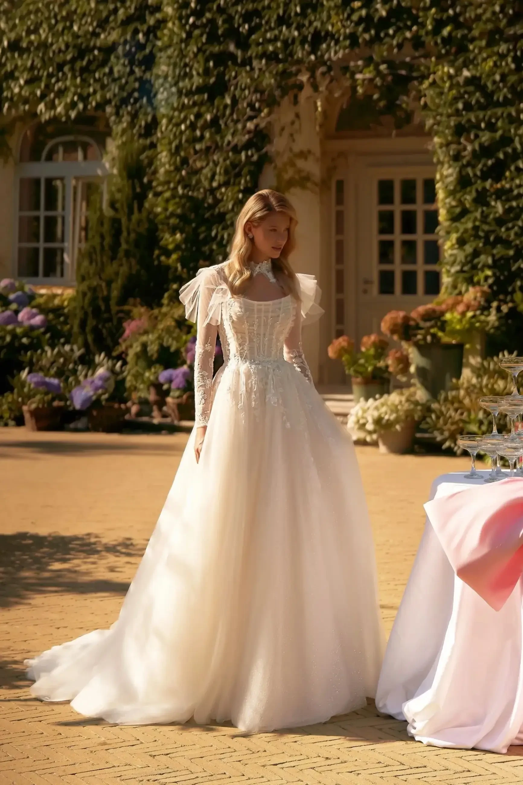 Model is wearing a soft A-line wedding gown with beaded straps, a pleated corset bodice, floral appliqués, and a layered glitter tulle skirt with a removable bolero.