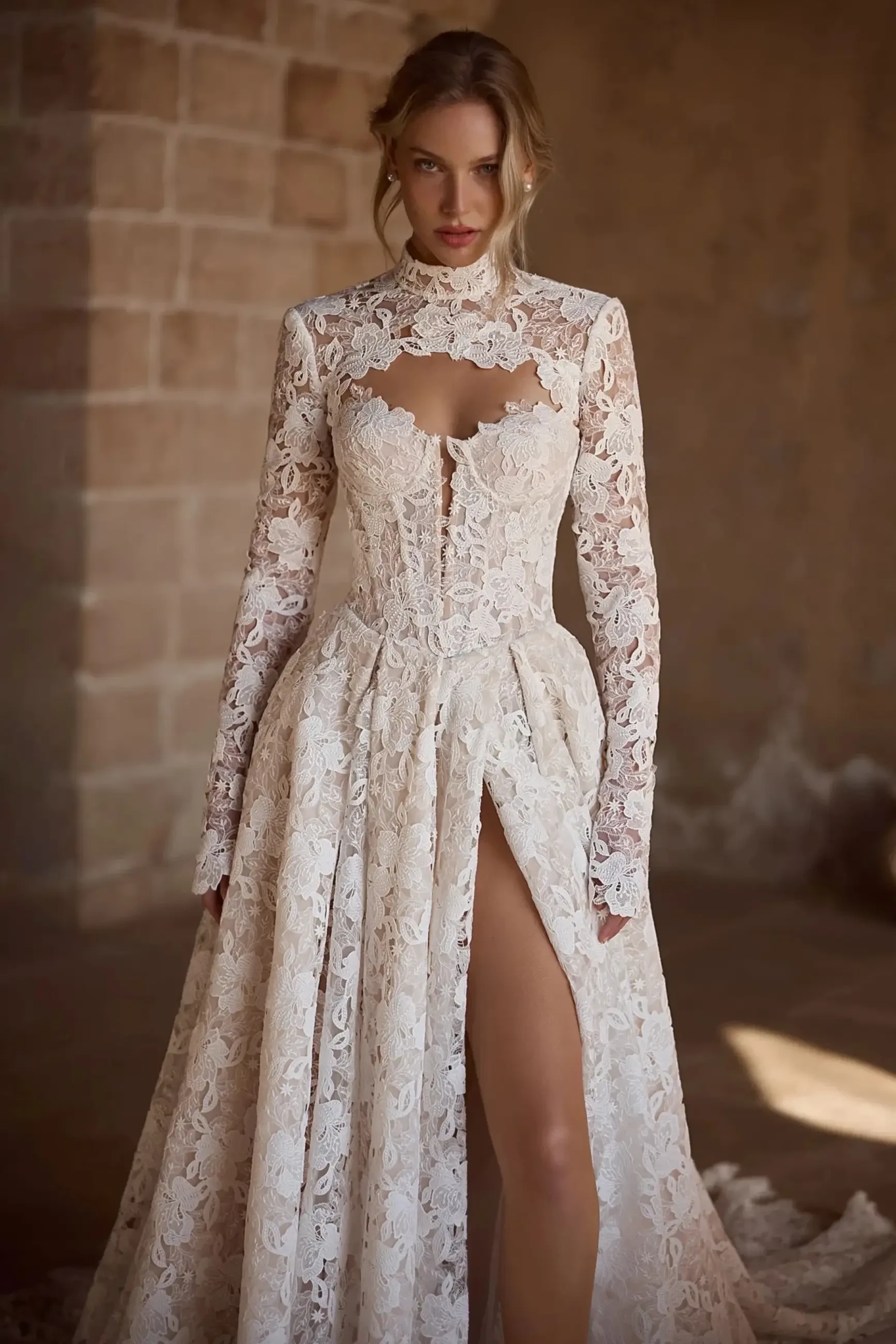 A model wearing a long-sleeve lace wedding dress with a high slit and intricate detailing. The dress features a fitted bodice and a flowing skirt, set against a textured wall backdrop.