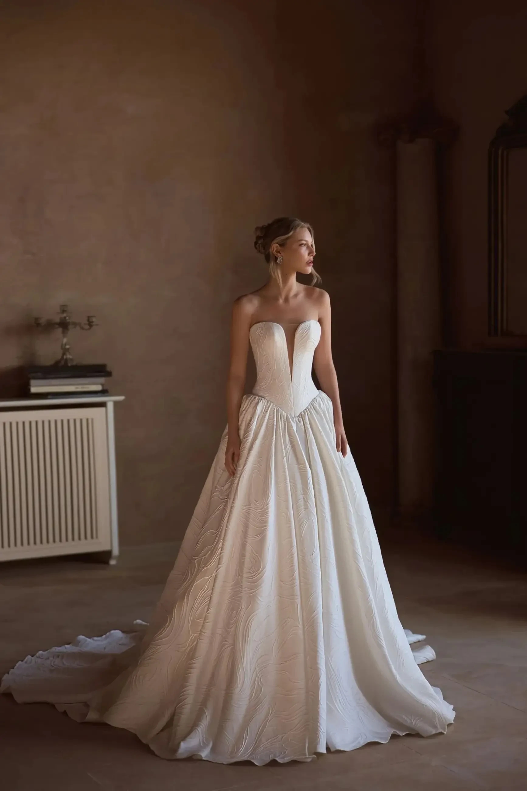 A woman in a strapless white wedding gown stands elegantly in a softly lit room, with a vintage decor and neutral tones.