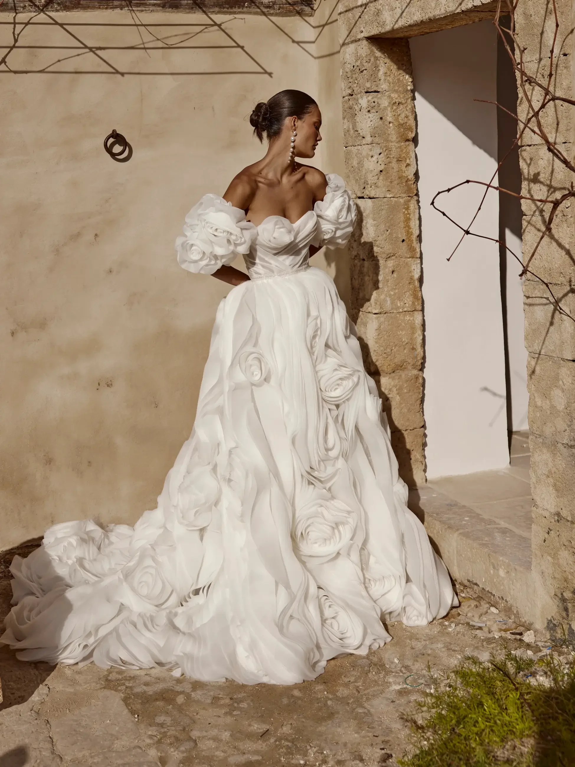 Model is wearing an off-the-shoulder organza gown with puffed sleeves, draped corset, a voluminous ruffled skirt resembling oversized roses, and a long trailing train.