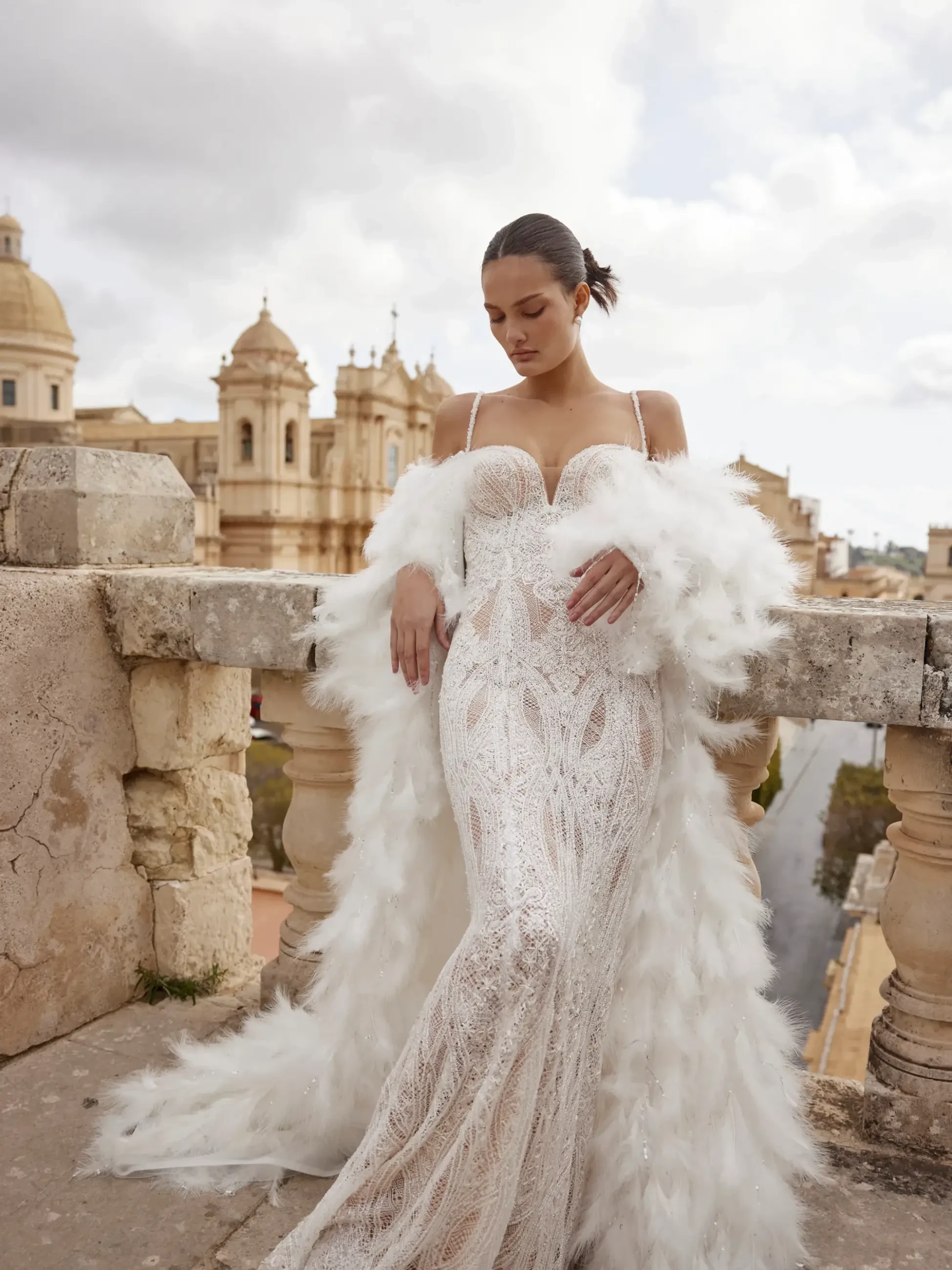 model is wearing a sleeveless sheath bridal gown paired with a floor length feather off the shoulder coat