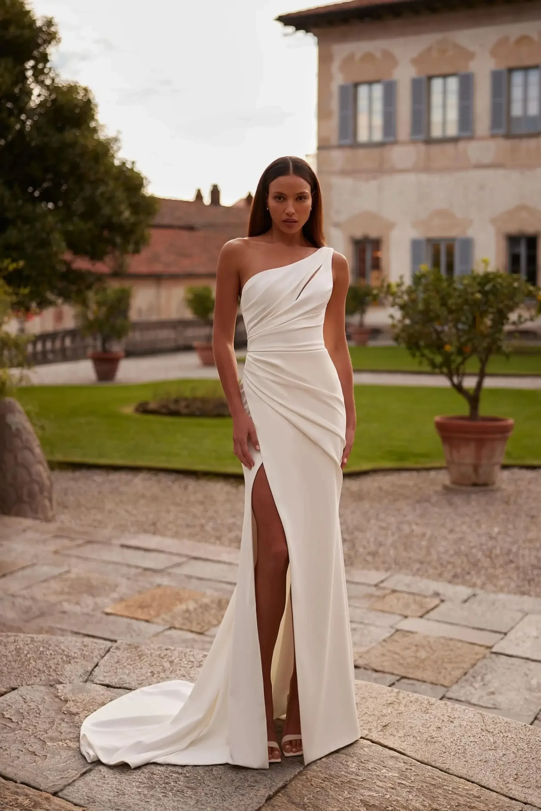 A woman in an elegant, one-shoulder white gown with a thigh-high slit stands confidently in a courtyard. The setting features lush greenery and a large historic building, creating a sophisticated and serene atmosphere.