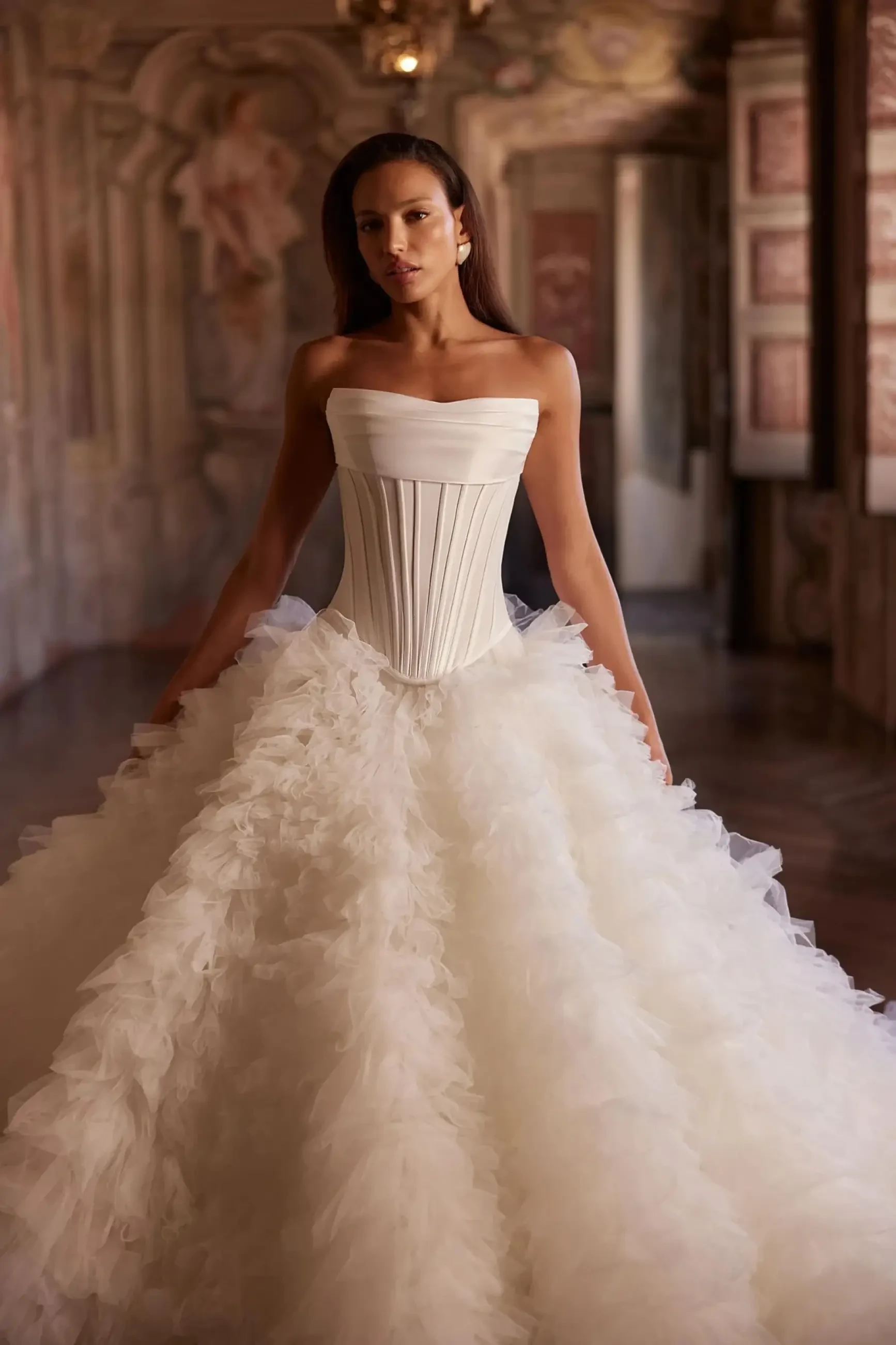 A woman in an elegant, strapless white gown with a structured bodice and a voluminous, textured skirt stands in an ornate, historic room.