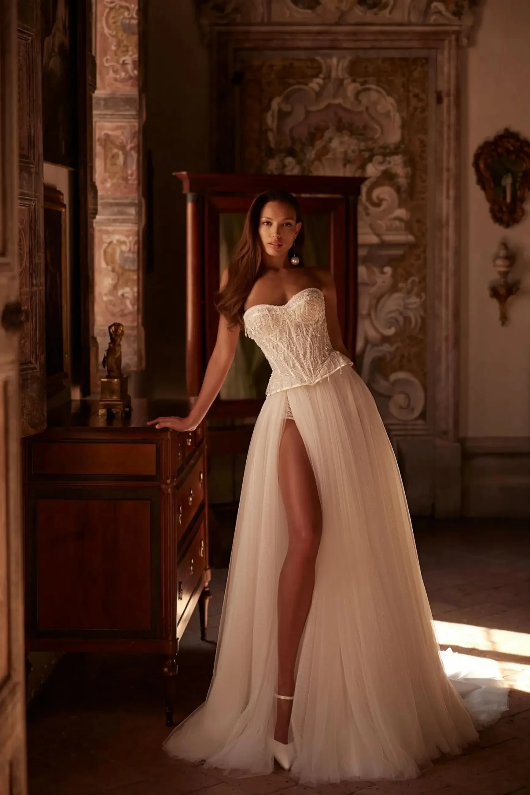 A woman in a flowing white gown with a thigh-high slit stands gracefully in an ornate room, leaning on an antique wooden table, exuding elegance and poise.