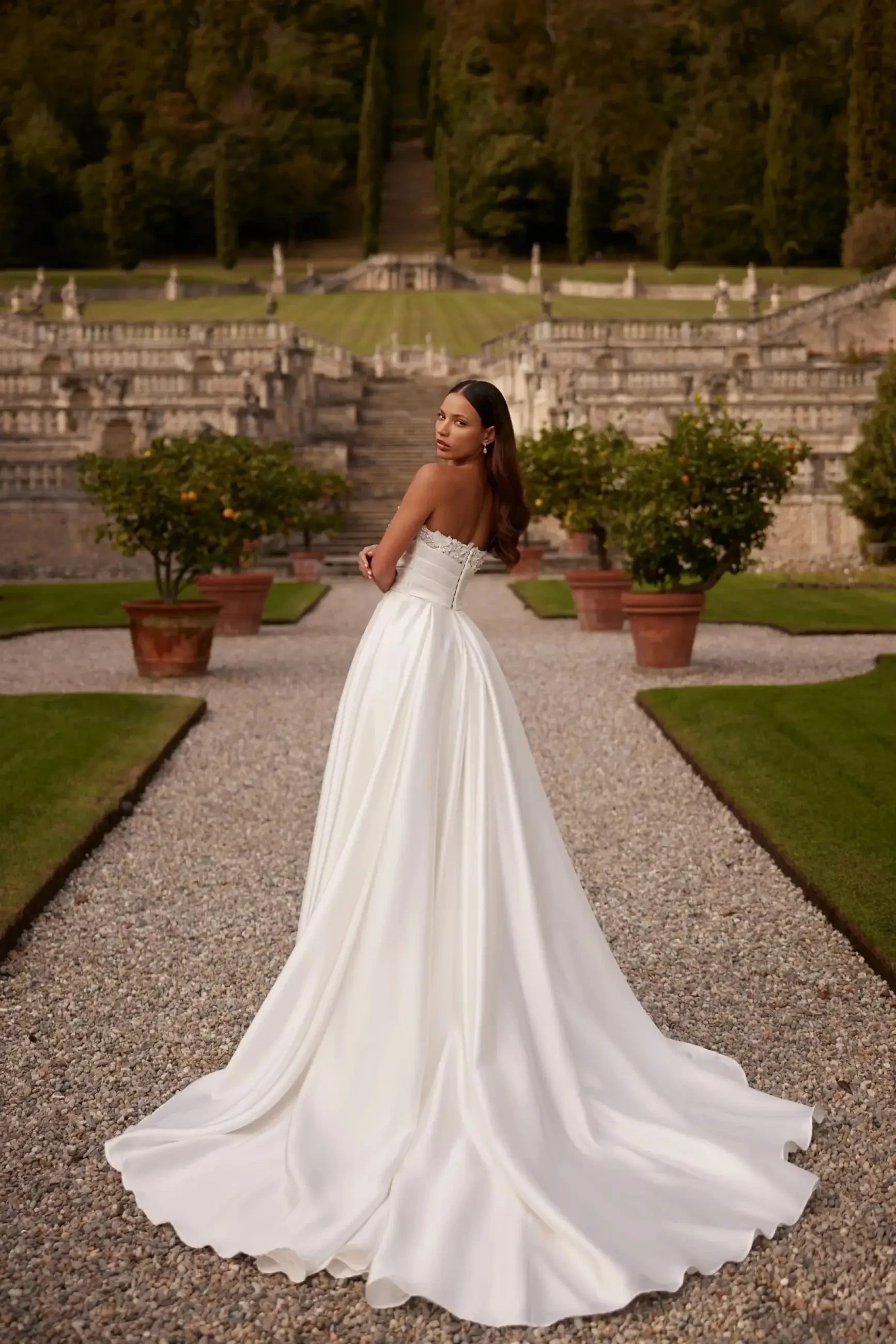 Model is wearing an ivory tulle gown with a semi-transparent lace corset, pearl-embroidered straps, v-neckline, v-back, and a full pleated skirt with lace appliqués and a long train.