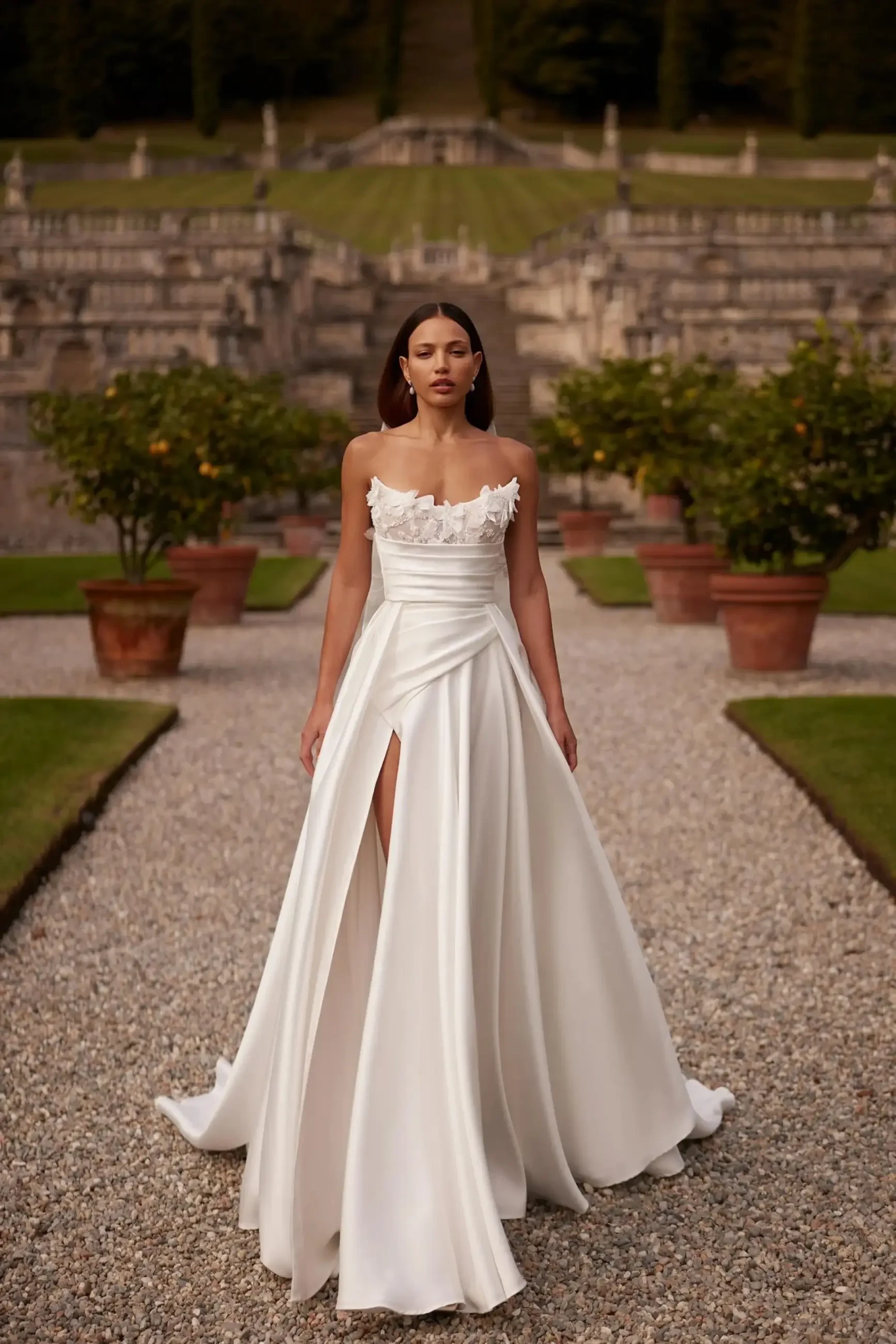 Model is wearing an ivory tulle gown with a semi-transparent lace corset, pearl-embroidered straps, v-neckline, v-back, and a full pleated skirt with lace appliqués and a long train.