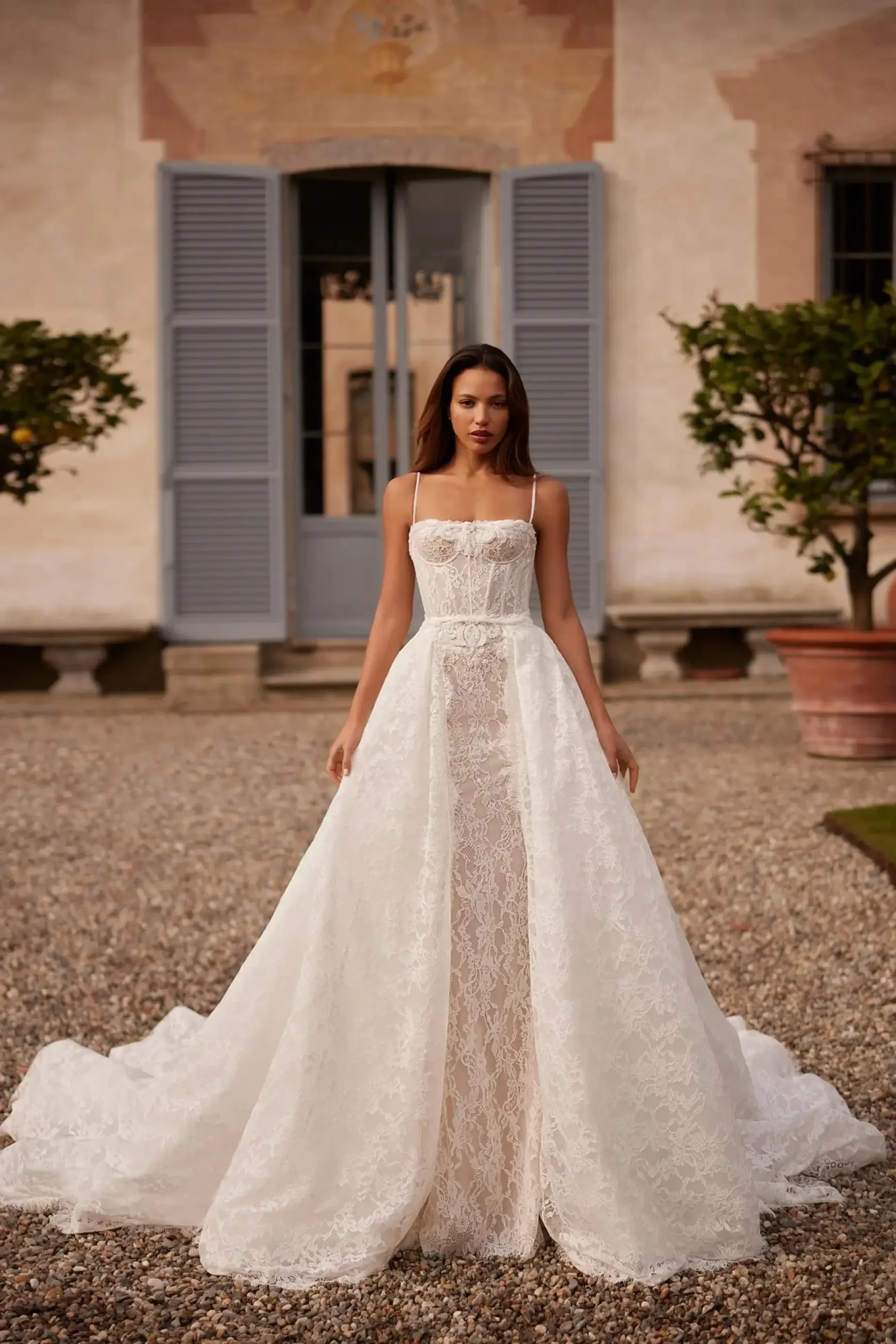 A woman stands elegantly in front of a rustic building in a flowing lace wedding gown with spaghetti straps. The background features blue shutters and potted plants, conveying a serene and timeless ambiance.