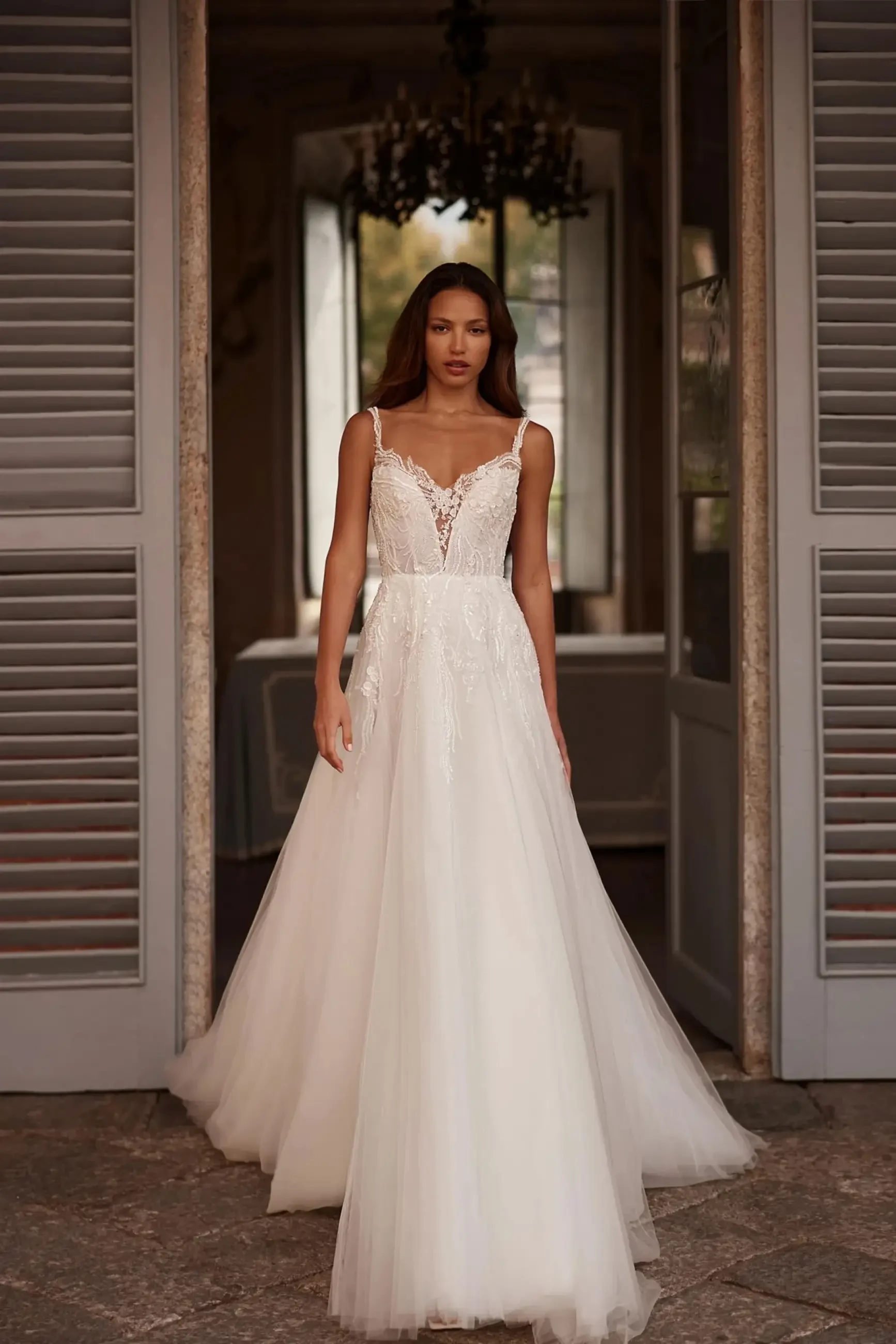 Woman in an elegant white lace wedding dress stands in a doorway with open shutters. She looks serene, with soft lighting highlighting the gown's details.