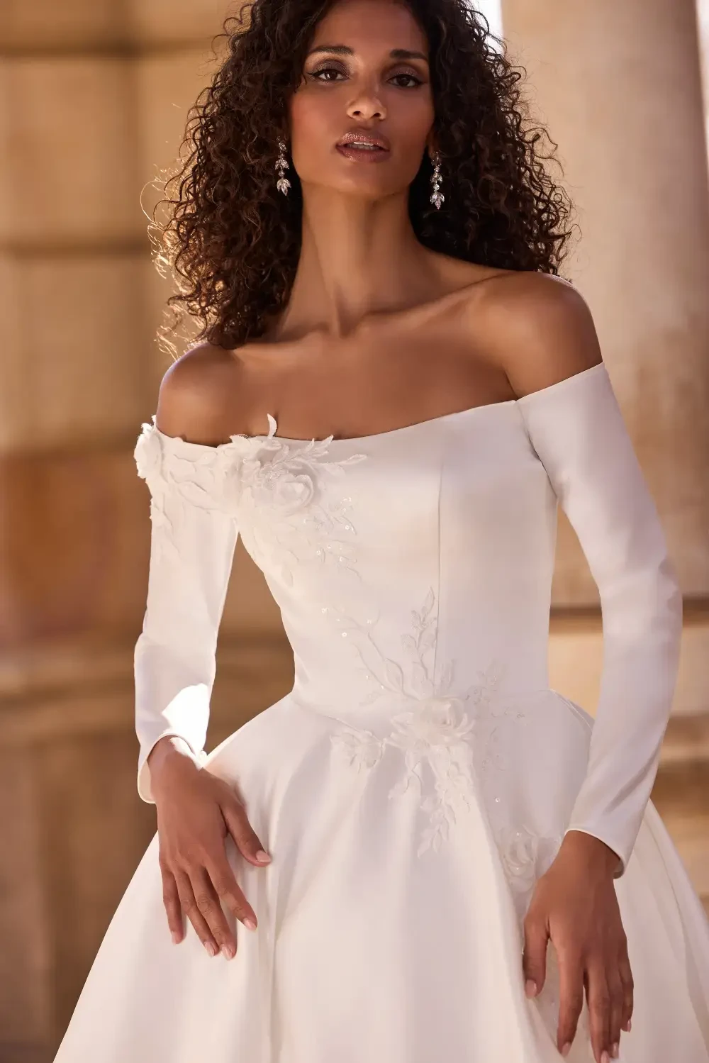 A bride wearing a white off-shoulder wedding dress with long sleeves, featuring floral detailing, standing in a grand setting.