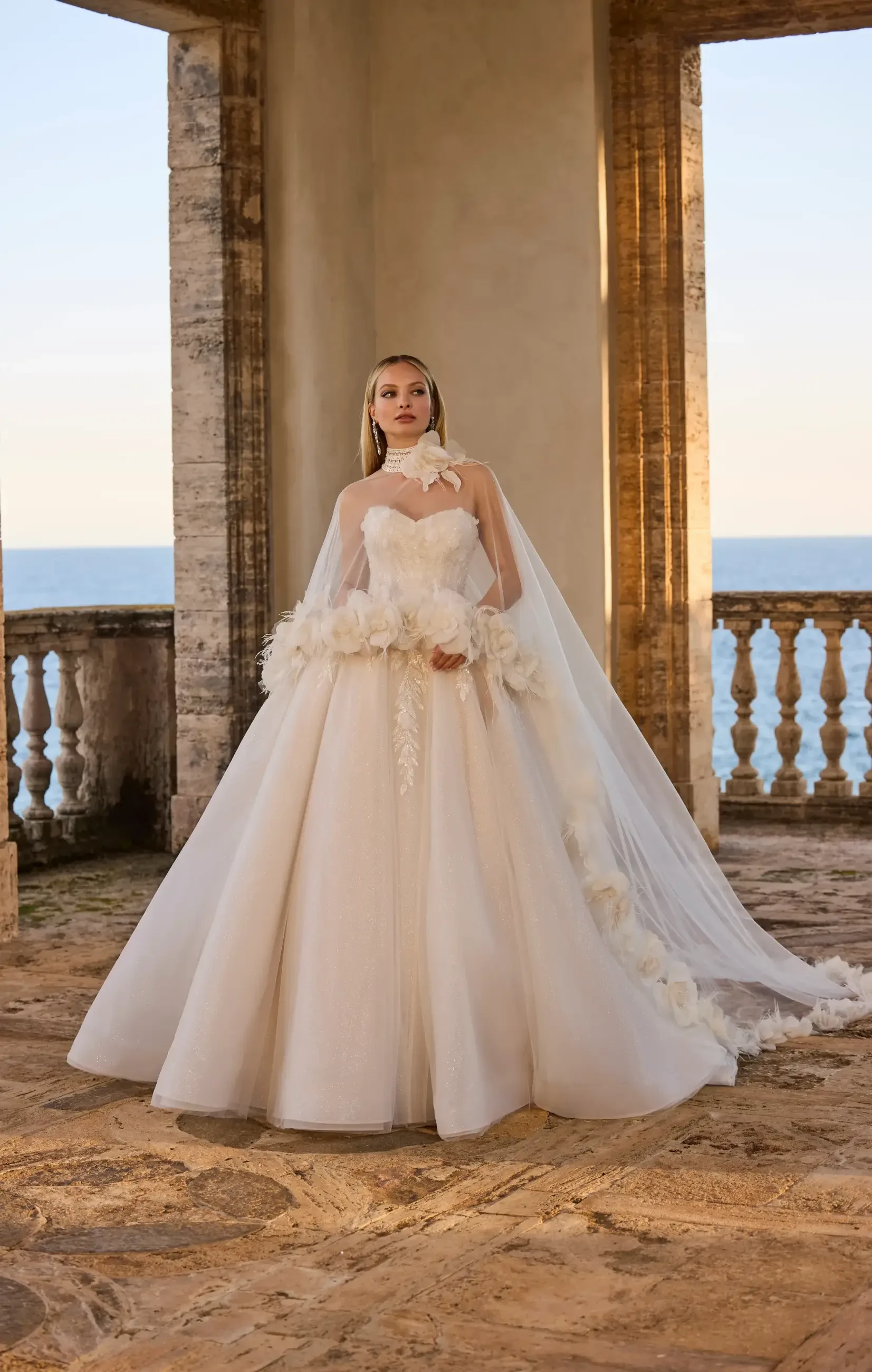 A woman in a strapless white wedding gown with floral details, standing gracefully indoors with a large window overlooking the sea in the background.