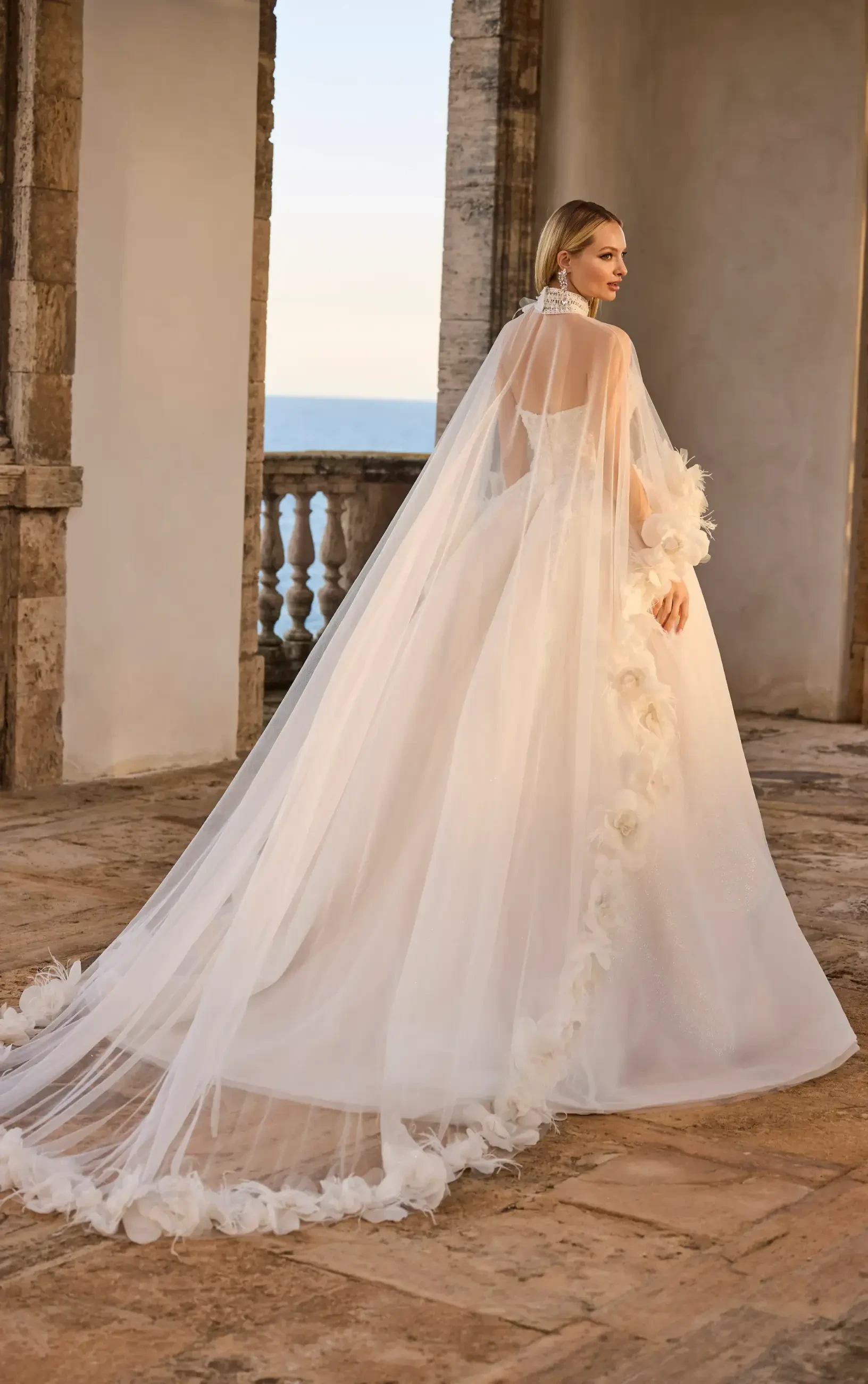 A bride in an elegant white gown with a long, flowing train adorned with floral details stands on a stone terrace, overlooking the ocean at sunset.