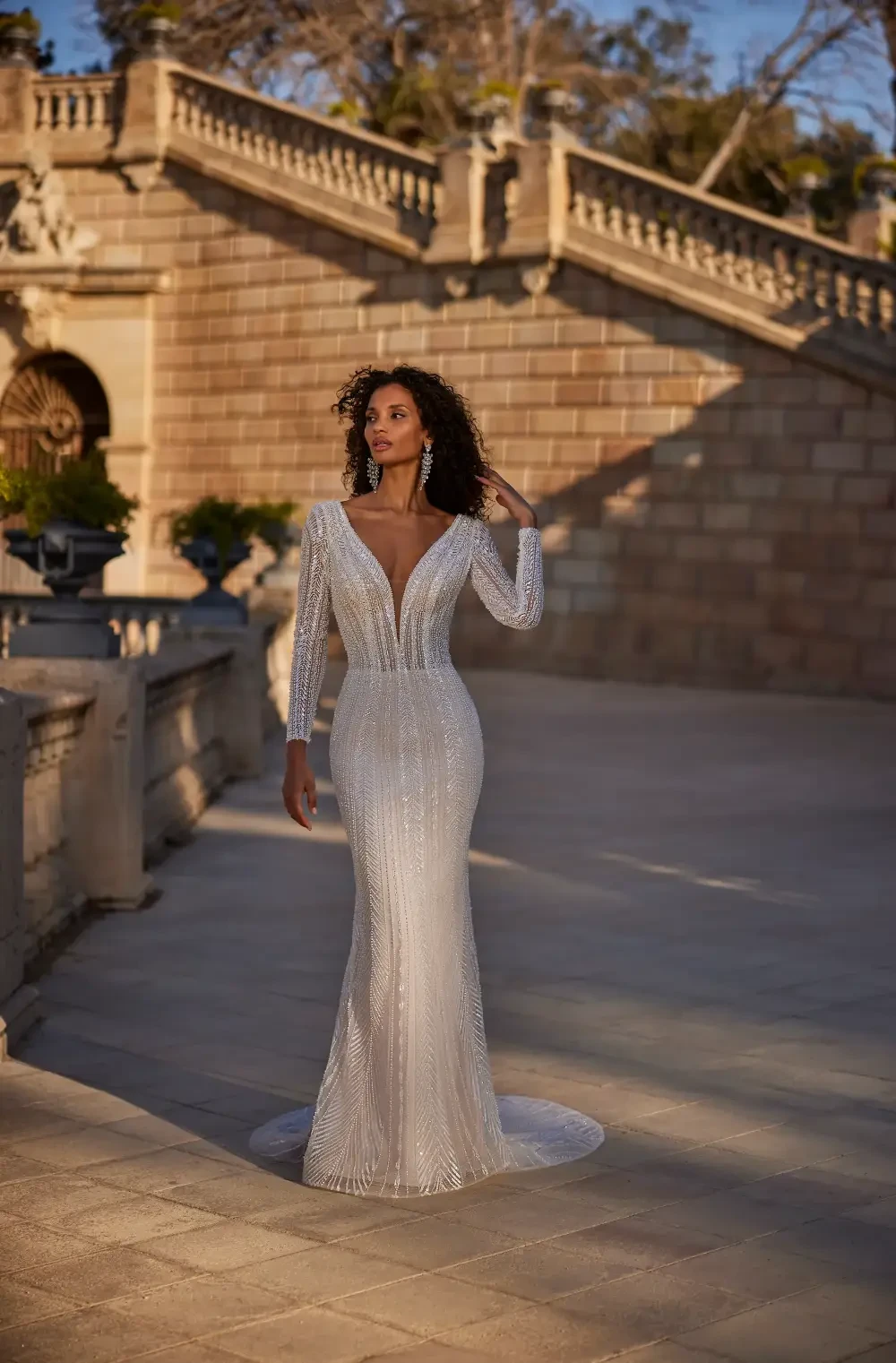 A woman in an elegant, shiny silver gown with long sleeves and a deep V-neck stands outdoors, posing gracefully in front of a grand architectural background.
