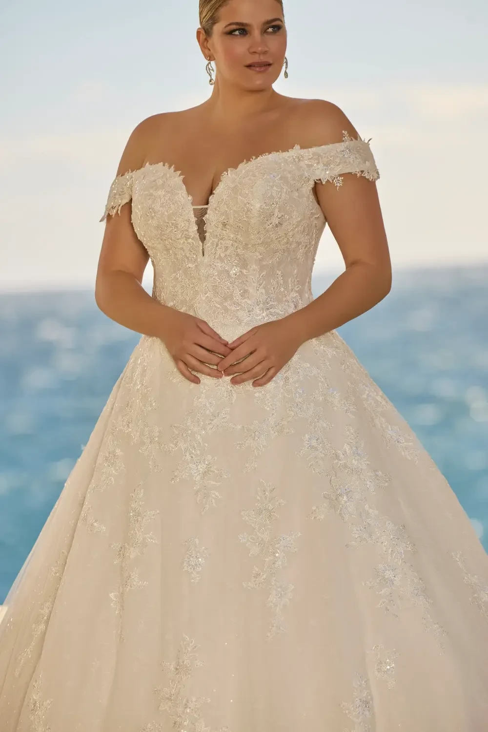 A woman wearing an off-the-shoulder wedding dress with intricate lace and bead details, standing in front of a blue ocean.