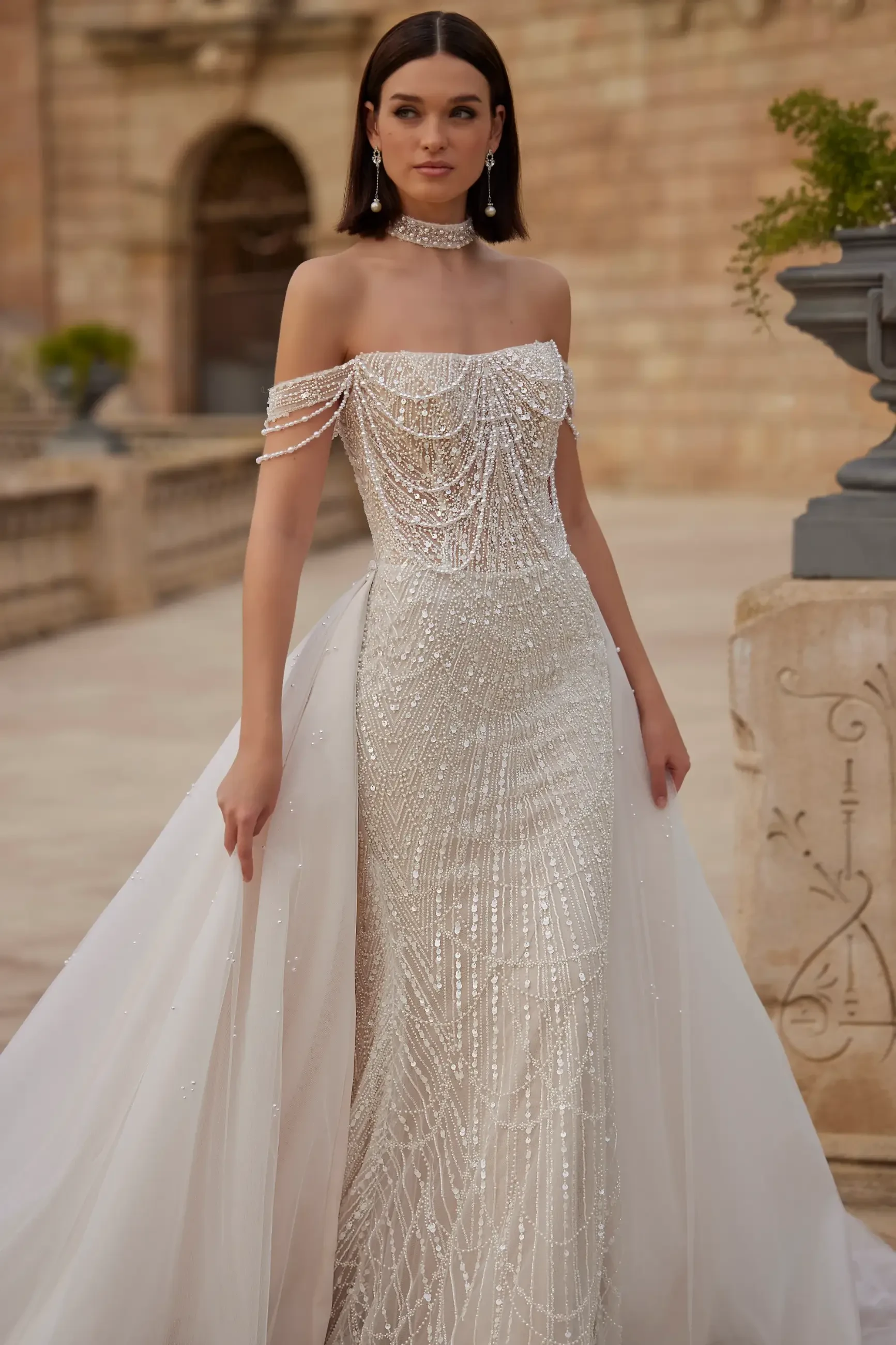 A woman in a glamorous, beaded off-shoulder gown with a sheer train stands outdoors against an ornate stone backdrop, exuding elegance and grace.