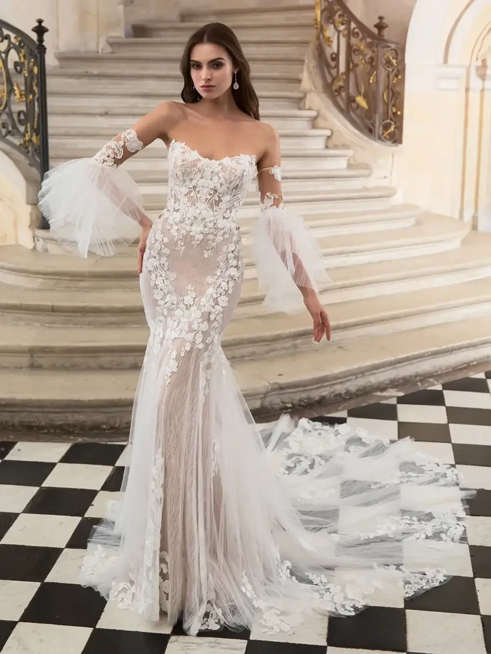 A model wearing a stunning lace wedding dress with floral detailing, off-the-shoulder sleeves, and a flowing train, posed on a checkered floor in an elegant interior setting.