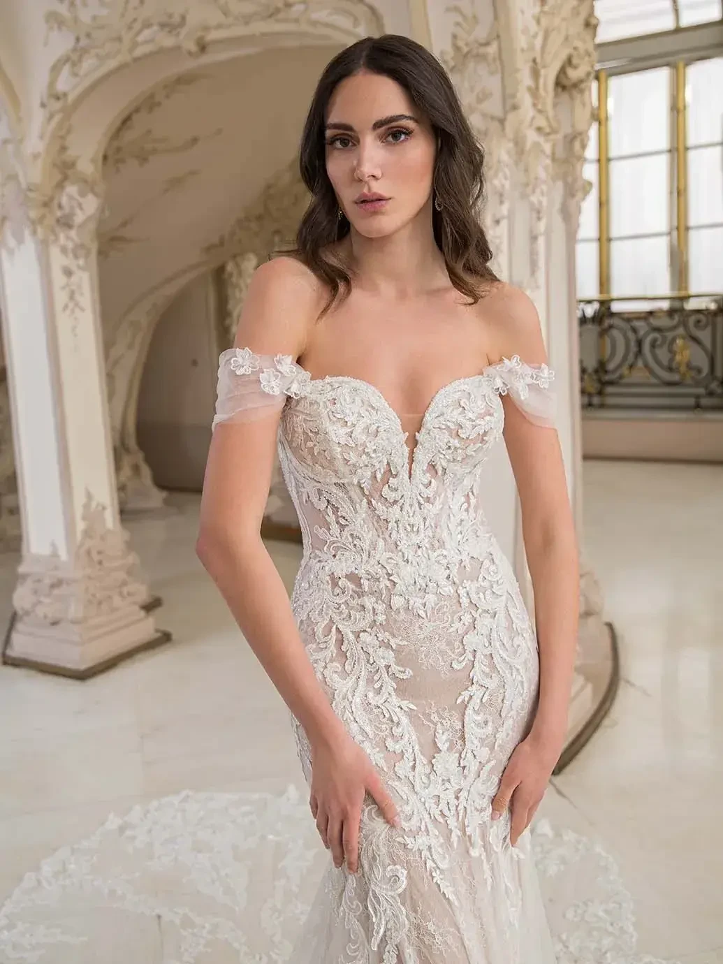 A woman in an elegant off-the-shoulder wedding dress with intricate lace detailing, standing in a beautifully ornate setting with large windows.