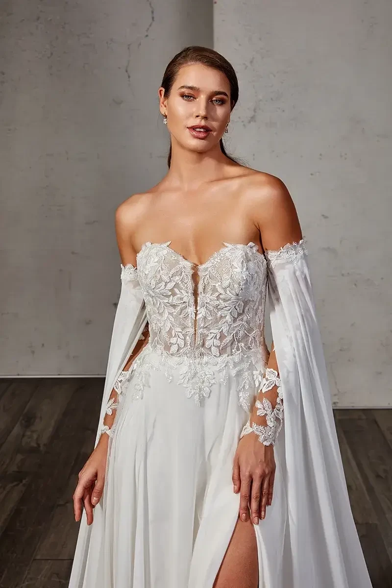 A model wearing an elegant white wedding dress with floral lace details and off-the-shoulder sleeves, posing in a minimalist setting with wooden flooring.