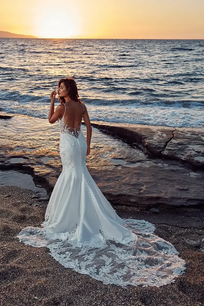 A woman in a backless white wedding dress stands on a beach at sunset, looking over her shoulder towards the ocean.