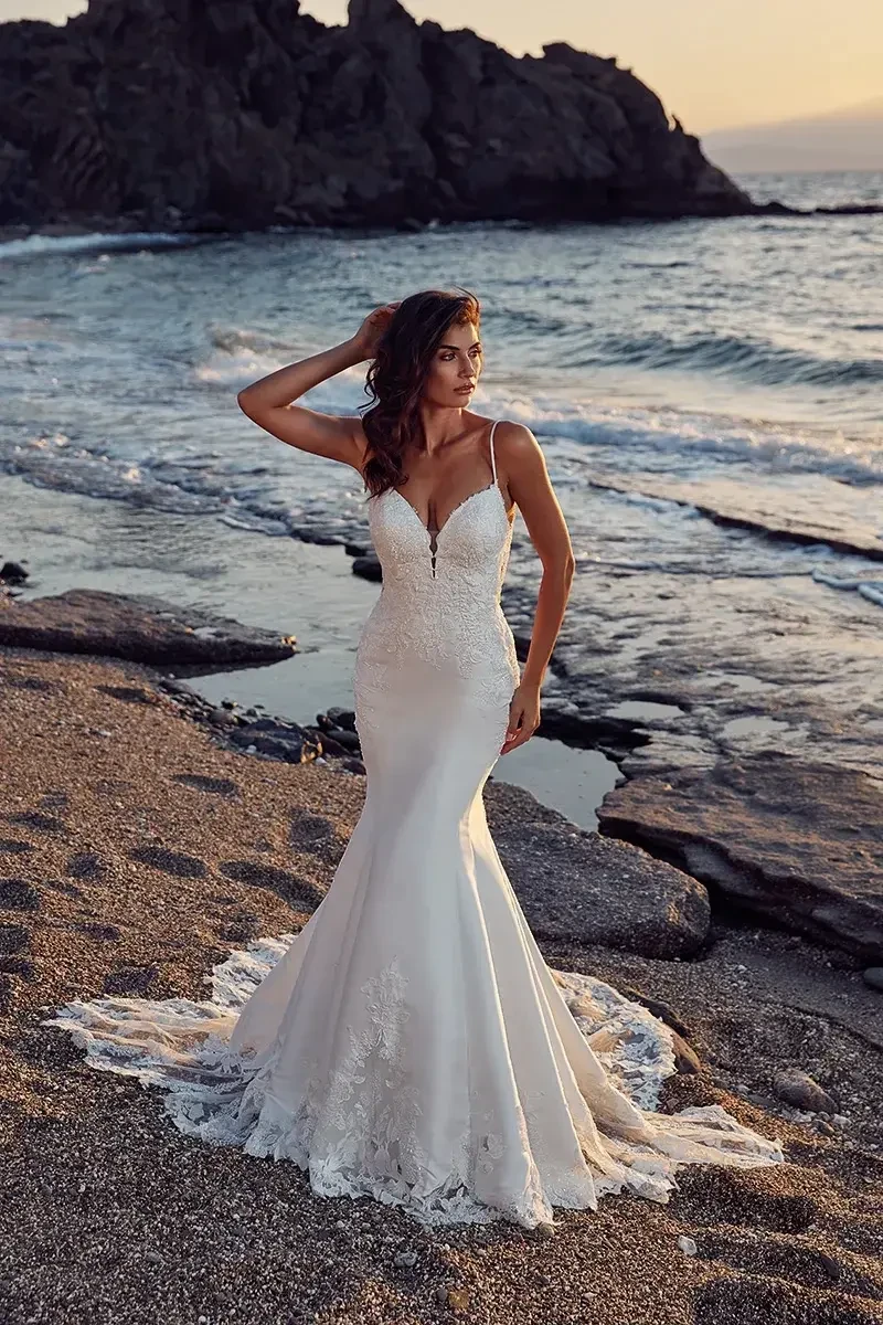 A woman in a fitted white wedding dress standing on a beach near the water, with rocks and waves in the background.