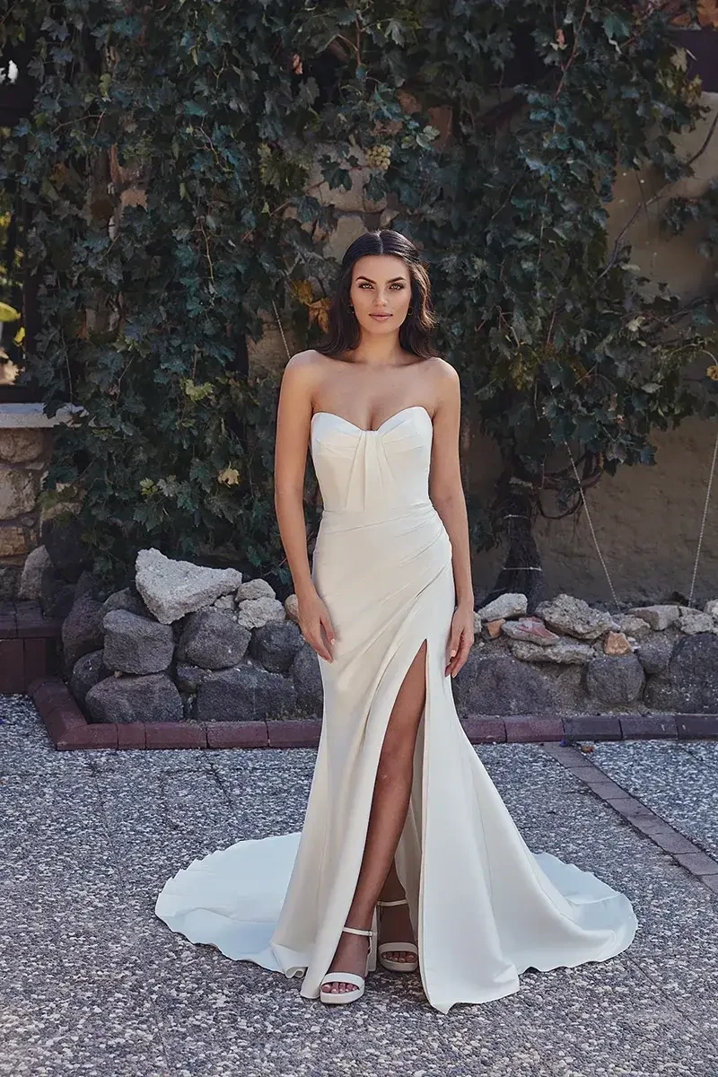 A woman in a form-fitting white gown with a thigh-high slit, standing on a gravel surface with greenery in the background.
