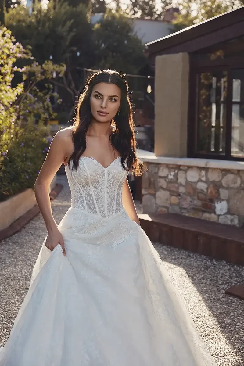 A woman in a strapless, corset-style wedding dress with a full skirt stands outdoors, looking towards the camera. The scene is set in a garden with trees and a stone building in the background.
