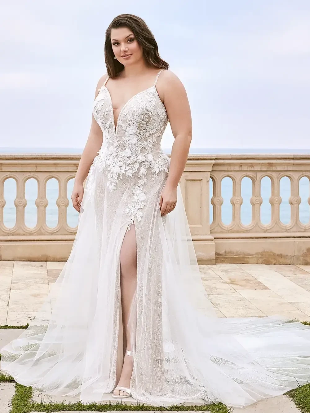 A woman in a beautiful, beaded white wedding gown with a deep V-neckline and a high slit, standing outdoors with a scenic background.