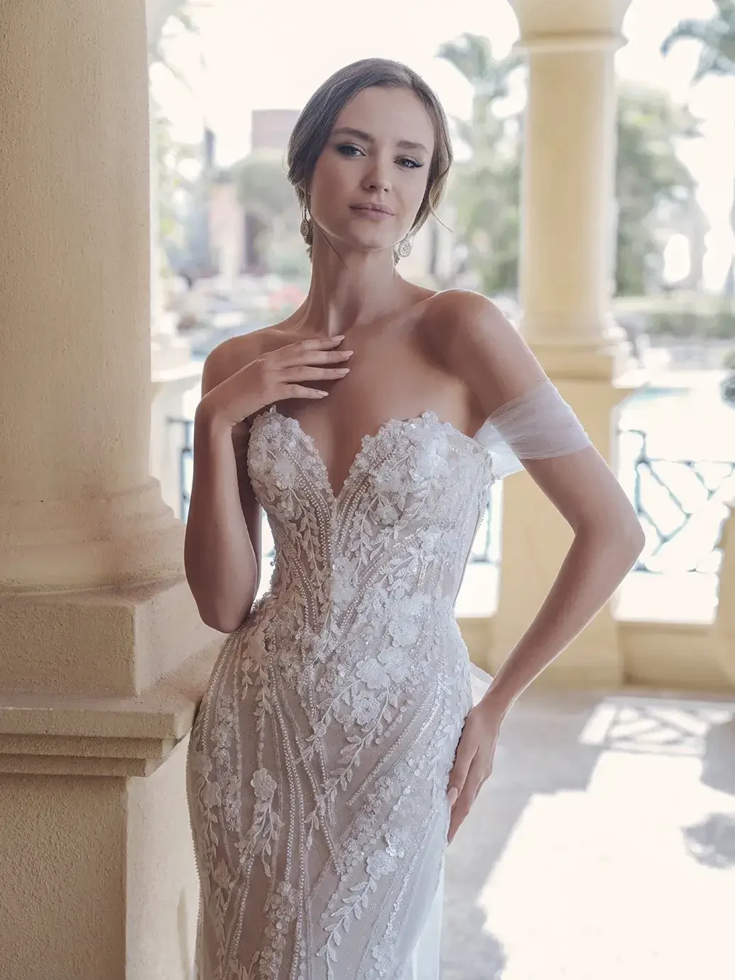 A woman wearing a beautiful lace wedding dress, standing gracefully by a large column with sunlight streaming in.