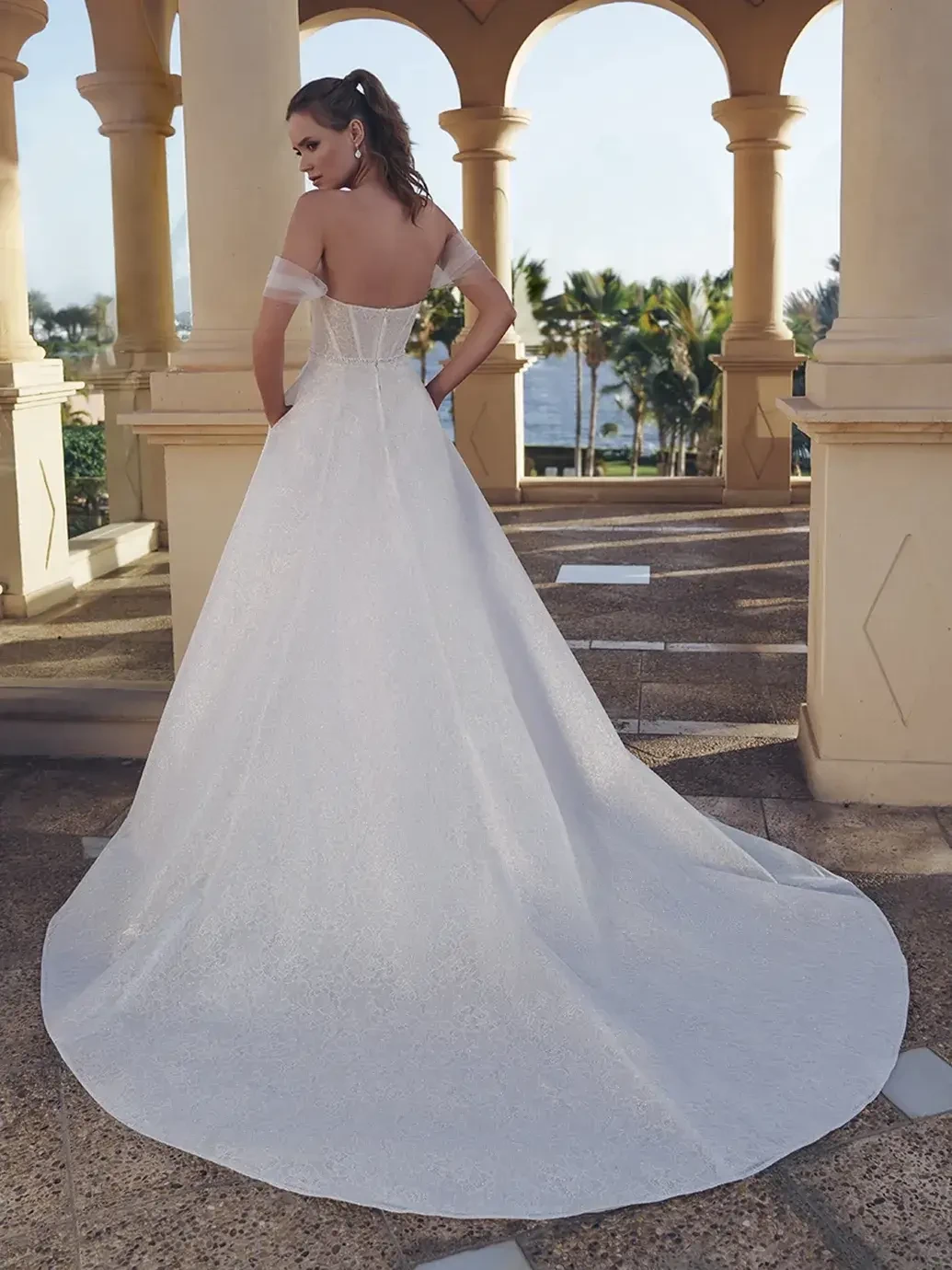 Model is wearing a glittering lace and organza A-line gown with a beaded waist belt, sweetheart neckline, detachable off-the-shoulder straps, and a front slit.