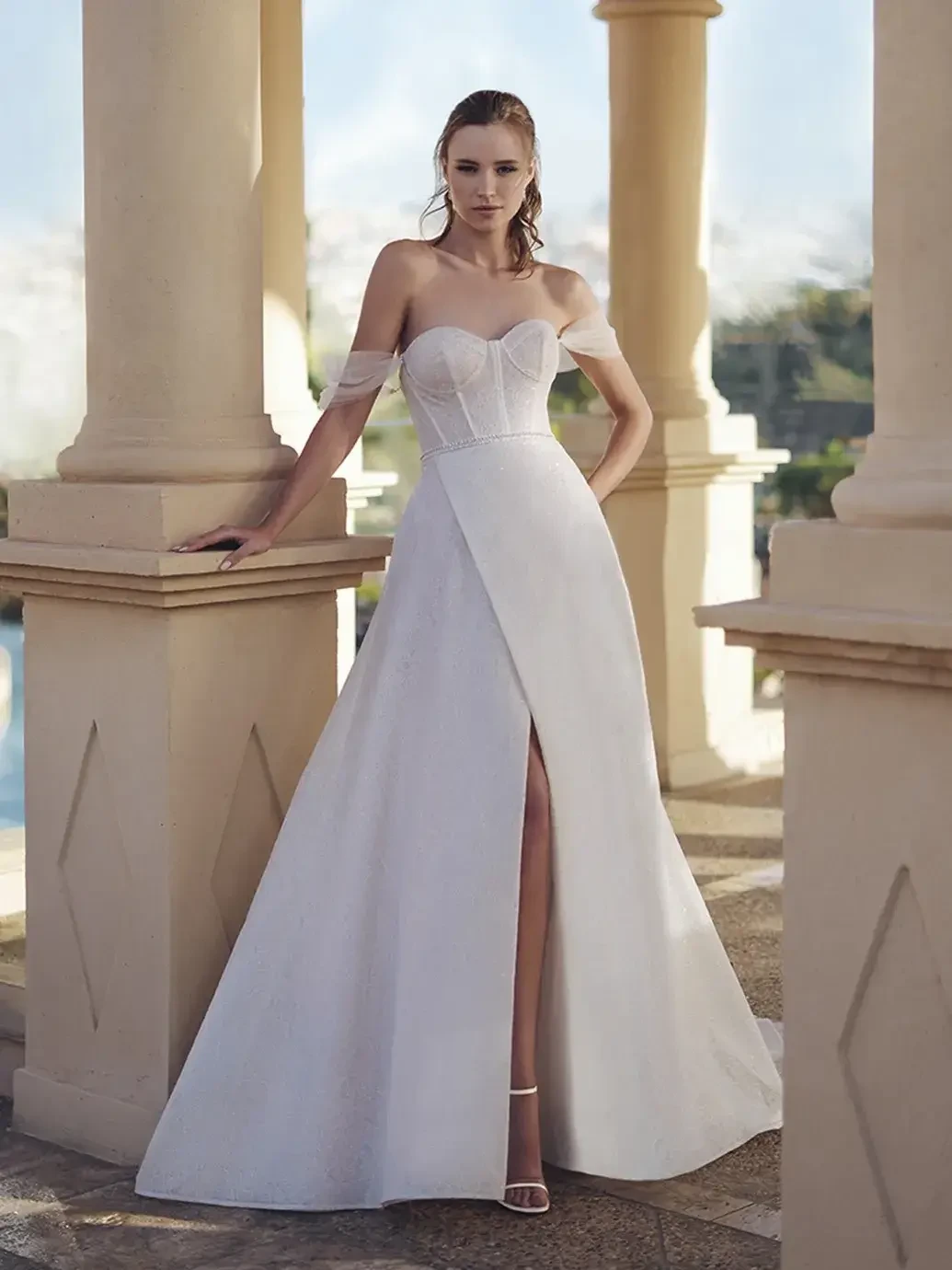 Model is wearing a glittering lace and organza A-line gown with a beaded waist belt, sweetheart neckline, detachable off-the-shoulder straps, and a front slit.