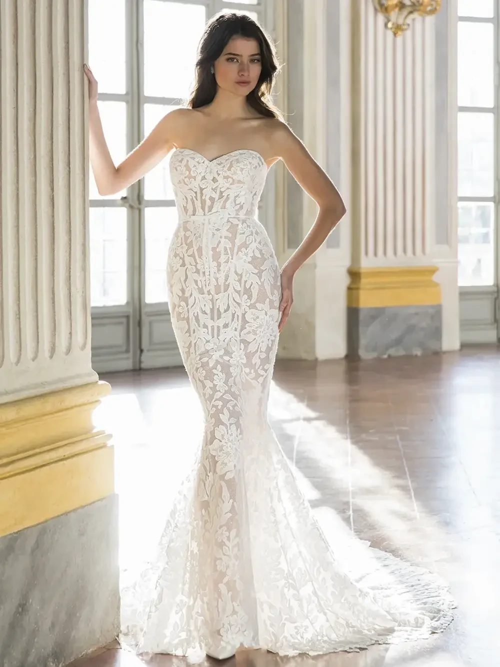 A model is wearing a strapless white wedding dress with a fitted silhouette and intricate lace details, posing in a bright, elegant indoor setting.