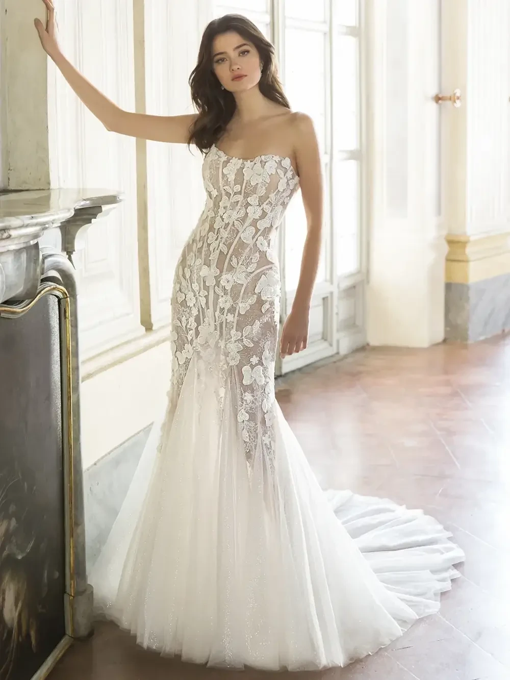 A model wearing a fitted, strapless wedding gown with intricate floral lace detailing and a flowing tulle train, posed in an elegant indoor setting.