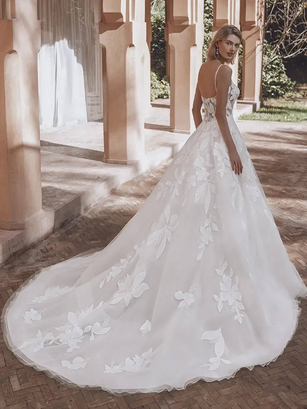 Model is wearing an A-line gown with a scoop neckline, 3D chiffon appliques, a structured illusion corset bodice, beaded spaghetti straps, and a flowing tulle and organza skirt with an illusion lace back.