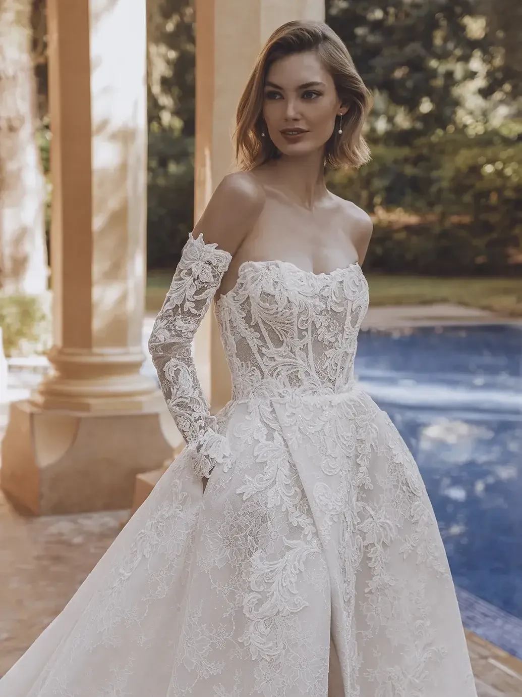 Model is wearing a ball gown with off the shoulder gauntlet sleeves with sequin floral lace